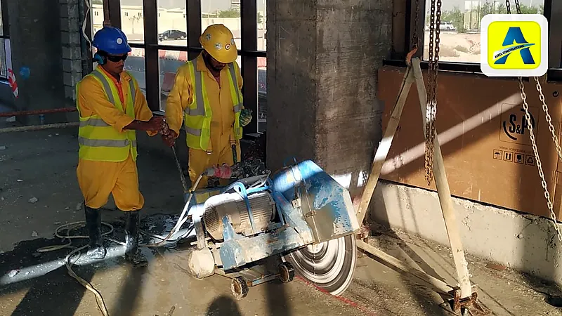 Two Aseel workers guide a floor saw through a concrete slab while a chain block hangs from a tripod, ready to lift the cut section out
